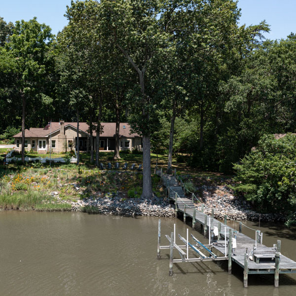 A dock in the foreground and in the background, up the bank is The Nest at Swan Point Virginia Airbnb vacation rental home, surrounded by trees.